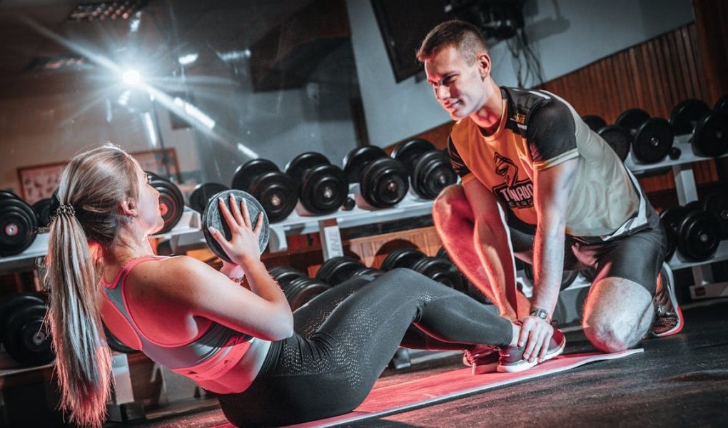 A personal trainer assists a woman doing sit-ups with a medicine ball in a gym setting.