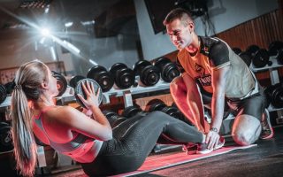A personal trainer assists a woman doing sit-ups with a medicine ball in a gym setting.